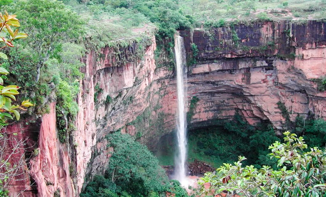 A tall, narrow waterfall cascades down a steep red rock cliff in Chapada dos Guimaraes, surrounded by lush green trees and vegetation, with mist forming at the base of the falls in a deep canyon.