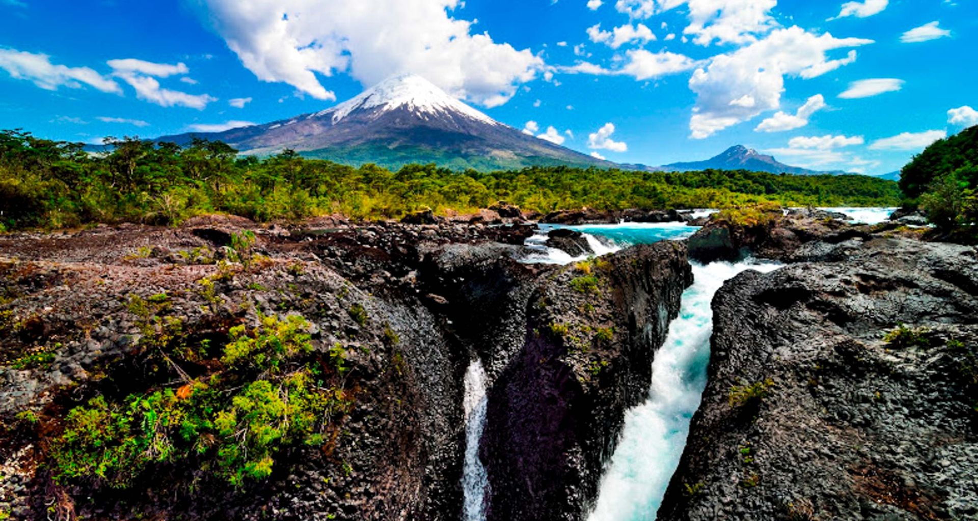A river with rapids flows through rocky terrain and lush greenery in Chile, with a snow-capped volcano amid the Andean Lakes, all under a bright blue sky with scattered clouds.