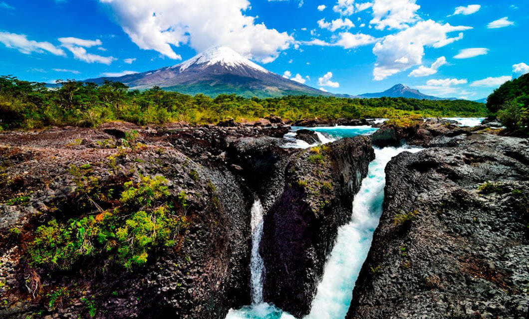 A river flows through dark volcanic rocks with lush greenery around in Chile, and a snow-capped volcano rises in the background under a bright blue sky with scattered clouds.