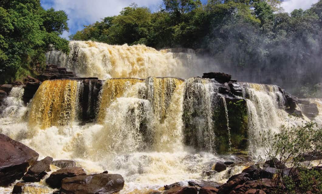 A powerful multi-tiered waterfall cascades over dark rocks, surrounded by lush green trees and mist—a vibrant scene that captures the beauty and force of one of the Venezuela highlights perfect for any Venezuela travel adventure.