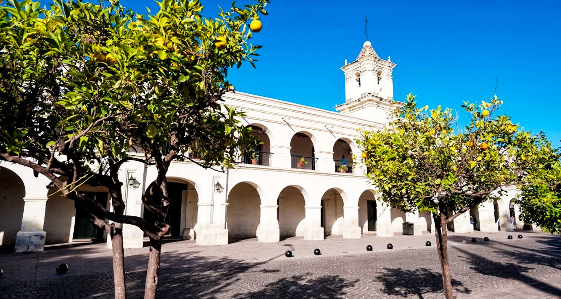 Colonial-style building with white arches and a bell tower in Salta, Argentina, seen behind orange trees in a sunny plaza with a clear blue sky—perfect for an Adventure Tour.