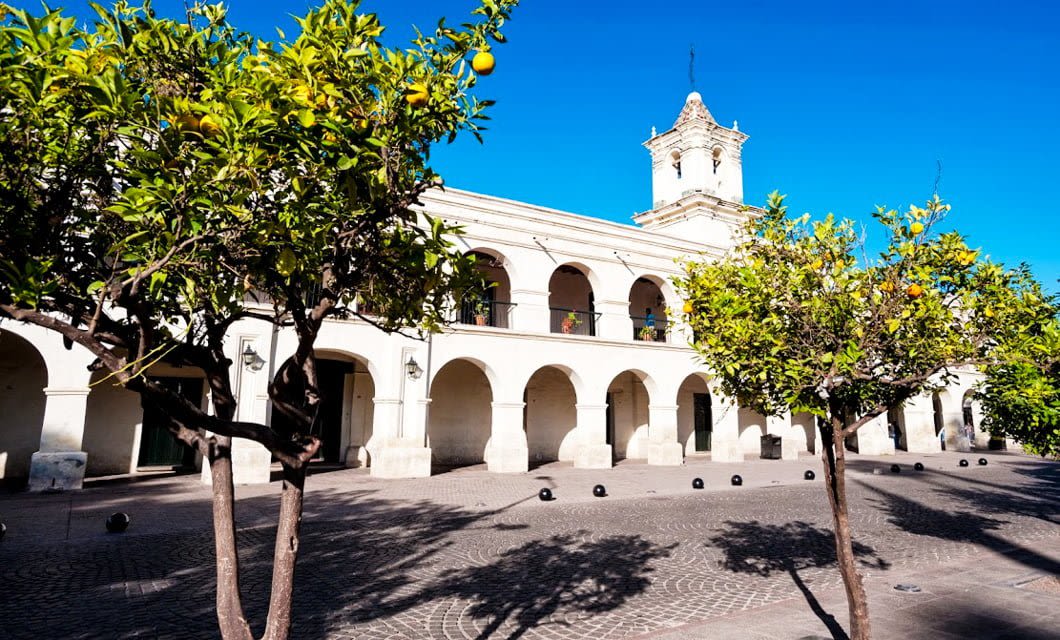 Historic white colonial building with arches and a bell tower in Salta, Argentina, surrounded by orange trees on a sunny day with a bright blue sky—perfect for an adventure tour.
