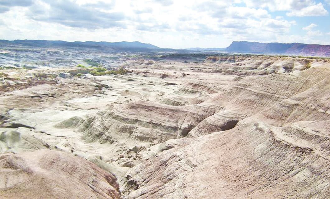 A wide, arid landscape with layered, eroded hills and rocky terrain under a partly cloudy sky, stretching toward distant mountains on the horizon—a timeless scene evoking the Boundaries of Time and the essence of a journey.