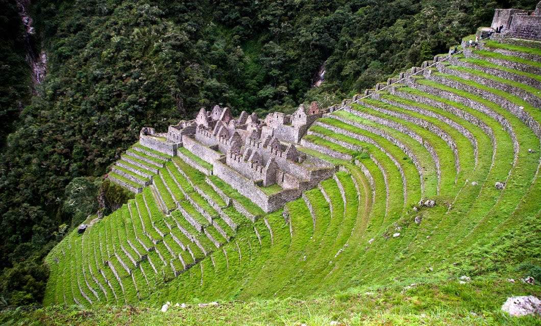 Ancient stone ruins sit among lush green agricultural terraces on a steep mountainside along the iconic Inca Trail to Machu Picchu, surrounded by dense forest. The terraces curve around the slope, showcasing impressive Incan engineering.