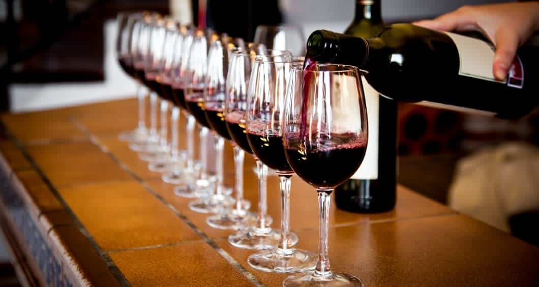 A person pours red wine into a row of glasses lined up on a tiled counter, evoking the charm of a railway journey like the famous Train to the Clouds, with filled glasses and wine bottles visible in the background.