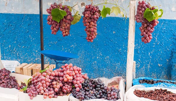 Large clusters of red and purple grapes hang above a market stall with bunches of grapes and other produce on a table, set against a blue and white wall—echoing the vibrant markets of the Sacred Valley. A blue weighing scale is also visible.