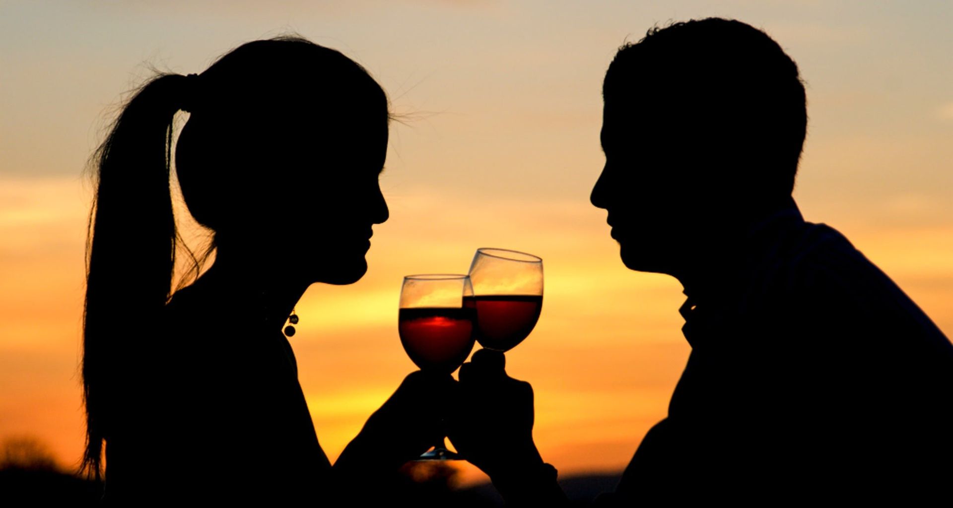 Silhouettes of a woman and man facing each other, clinking wine glasses at sunset. The orange and yellow sky creates a romantic atmosphere—perfect for a honeymoon in Mendoza.