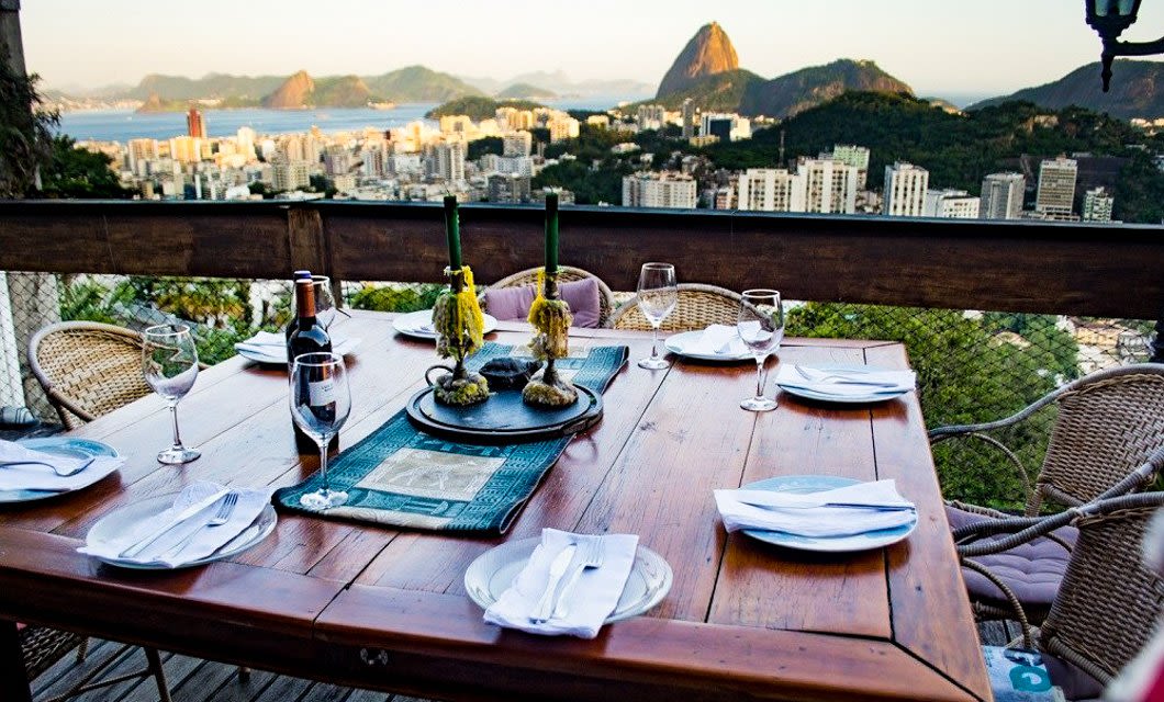 A wooden outdoor dining table set for six overlooks a cityscape with mountains and Sugarloaf Mountain in the distance. Wine glasses, plates, and candles are arranged for a delightful evening of Brazilian cuisine.