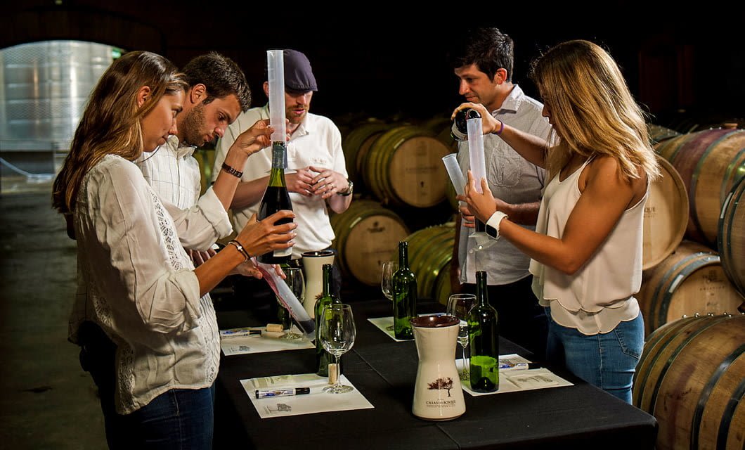 Five people stand around a table in a winery, each measuring wine in graduated cylinders with bottles, glasses, and papers nearby; wooden barrels are seen in the background—a perfect post-round scene after a day on the Chile Golf Tour.