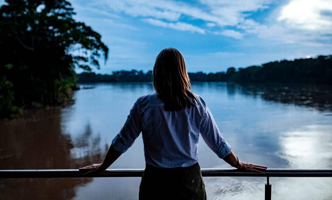A person in a white shirt stands on a balcony overlooking a wide river at dusk—perhaps dreaming of Anakonda Amazon Cruises—with trees lining the opposite bank and a cloudy blue sky above.
