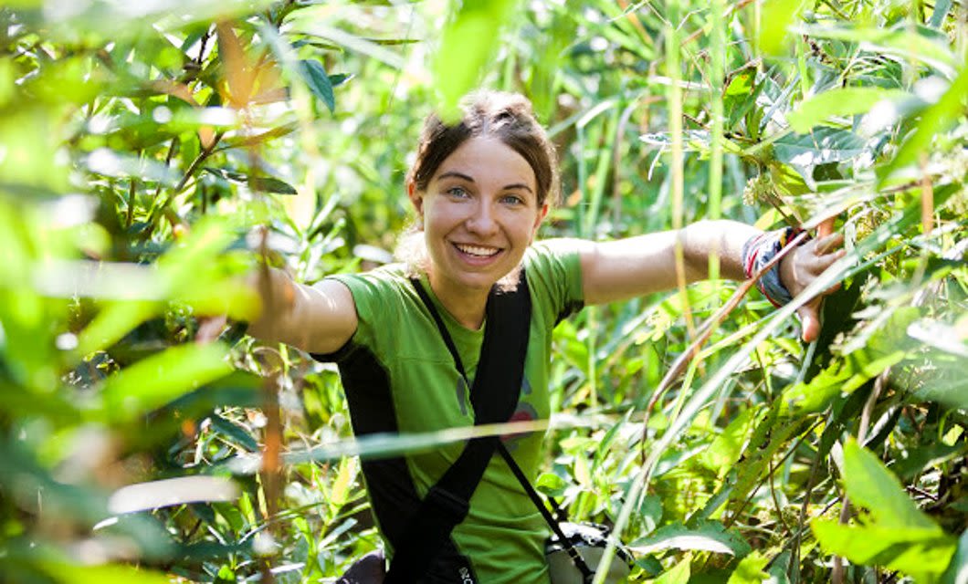A smiling woman wearing a green shirt and backpack pushes through dense, sunlit greenery in the Pantanal, surrounded by tall plants and leaves, enjoying an outdoor adventure.
