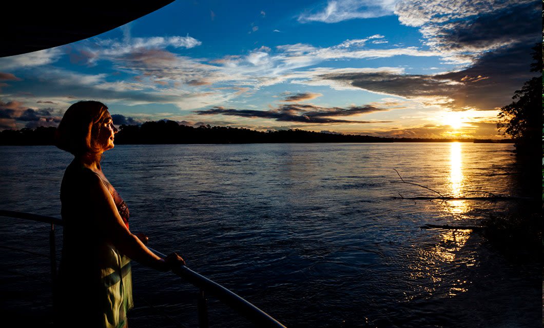 A person stands on a boat deck during a Manatee Amazon Explorer cruise, gazing at a vibrant sunset over a wide river in the Ecuadorian Rainforest, with dramatic clouds and shimmering reflections on the water.