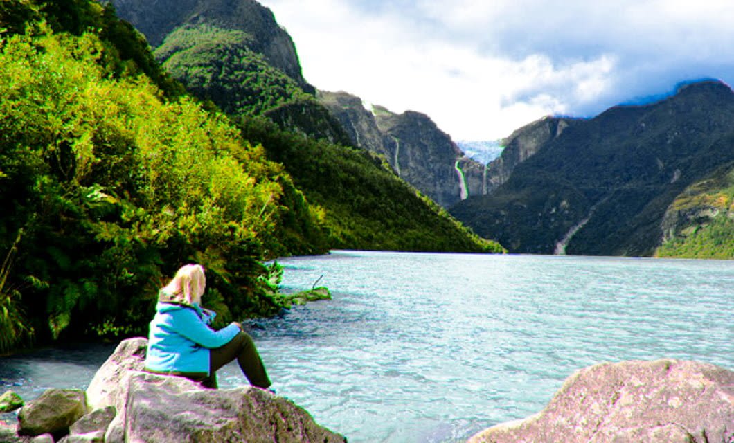 A person in a blue jacket sits on rocks by a calm lake in Northern Patagonia, surrounded by green mountains with a waterfall and cloudy sky in the background.