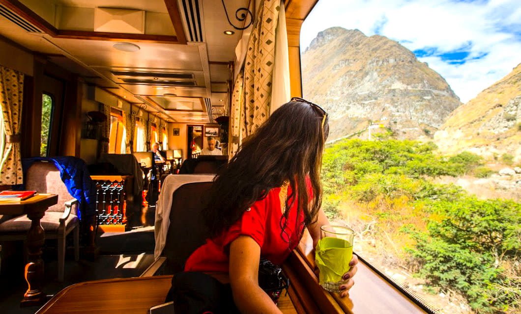 A woman in a red shirt holds a green drink and looks out the window of an Ecuador Trains ride at mountains and greenery under a bright sky. The train’s interior appears comfortable, with large windows and yellow curtains.