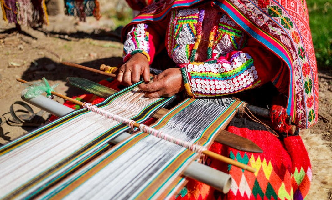 A person in colorful traditional clothing weaves fabric on a loom at an Amazon Lodge, using their hands to arrange threads. The vibrant patterns and detailed embroidery stand out in the sunlight.