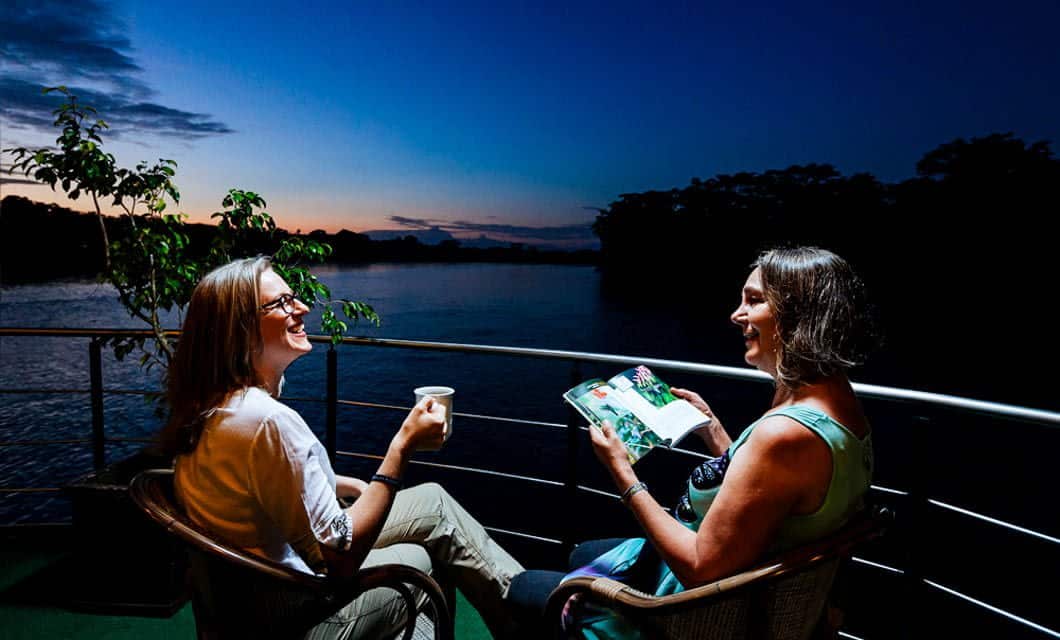 Two women sit on a balcony overlooking the river at sunset during their Anakonda Amazon Cruise. Smiling, one holds a cup and the other a magazine, enjoying the evening with trees and a colorful sky in the background.