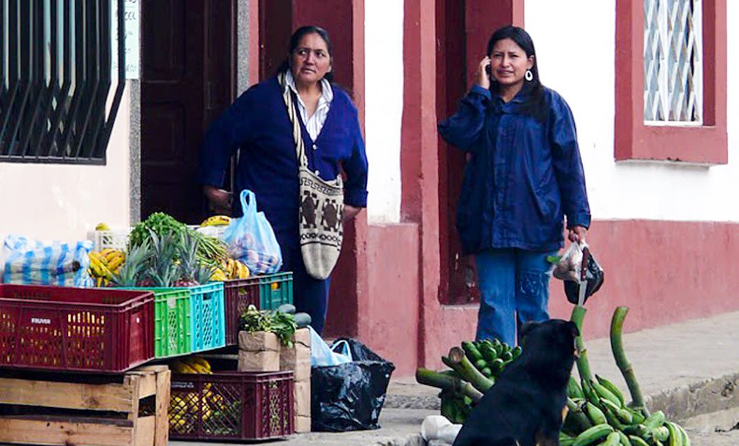 Two women stand on a sidewalk near crates of fruits and vegetables, evoking the vibrant street scenes often found during an Archaeology Tour. One talks on her cellphone while the other glances aside; a dog rests near long green produce on the ground.