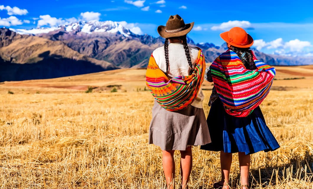Two women in traditional colorful clothing and hats stand in a dry field along the Lares Trek, facing distant snow-capped mountains under a bright blue sky with clouds.