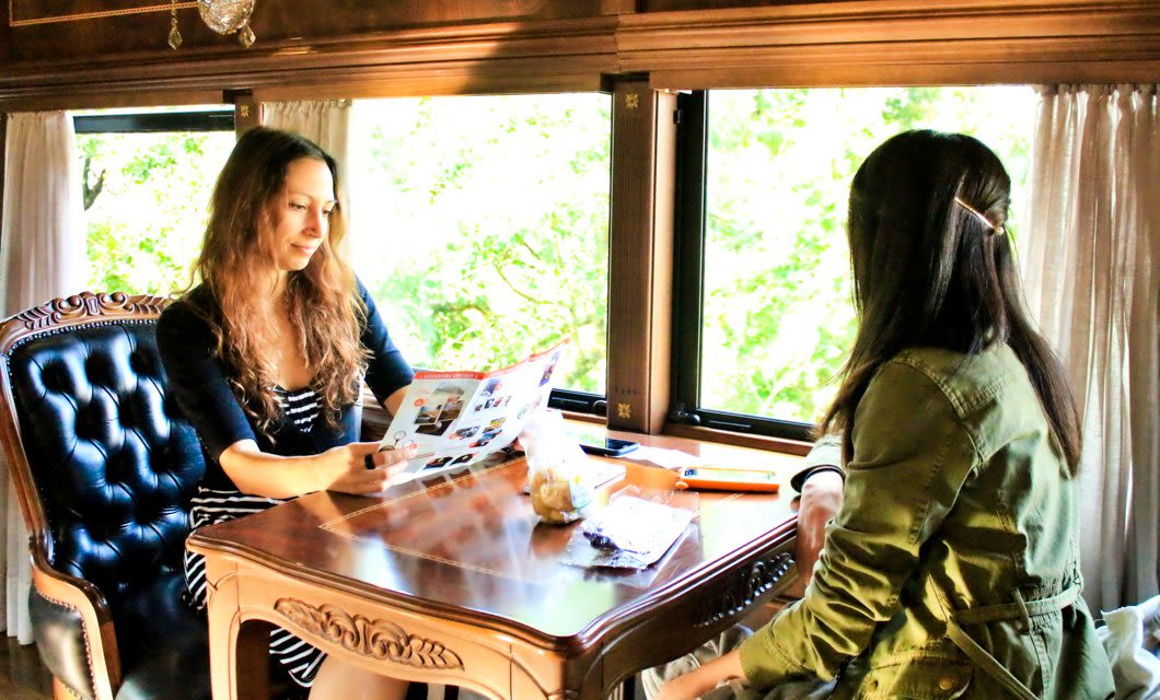 Two women sit at a wooden table by large windows, discussing a Brazil Rail Tour brochure. As one reads about the Historic Gold Route, the other listens. Papers and snacks are on the table, with greenery visible outside.