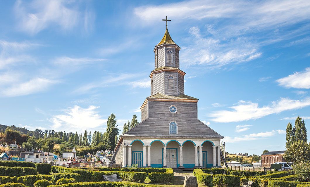 A wooden church with a tall steeple and cross stands under a blue sky along the Carretera Austral, surrounded by trimmed green bushes and trees, with a cemetery visible to the left.