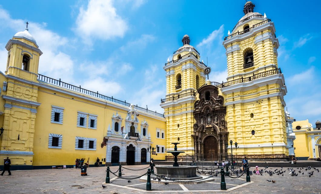 A large yellow colonial church with two tall towers and intricate brown stone detailing stands in a spacious plaza with a central fountain, evoking the charm of South America, surrounded by people and pigeons under a bright blue sky.