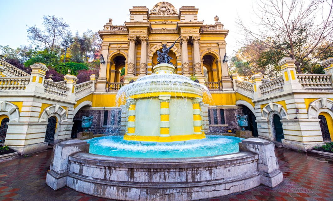A grand yellow and white fountain with a statue at the top stands before an ornate, historic building with arches and columns in Santiago, surrounded by trees and graceful staircases.