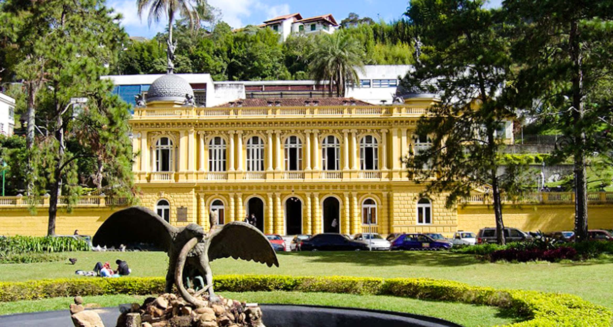 A large yellow neoclassical building with arched windows stands behind a park with green lawns, trees, and a fountain featuring an eagle statue—a picturesque spot often admired by those on a Brazil train tour like the famous Maria Fumaca.