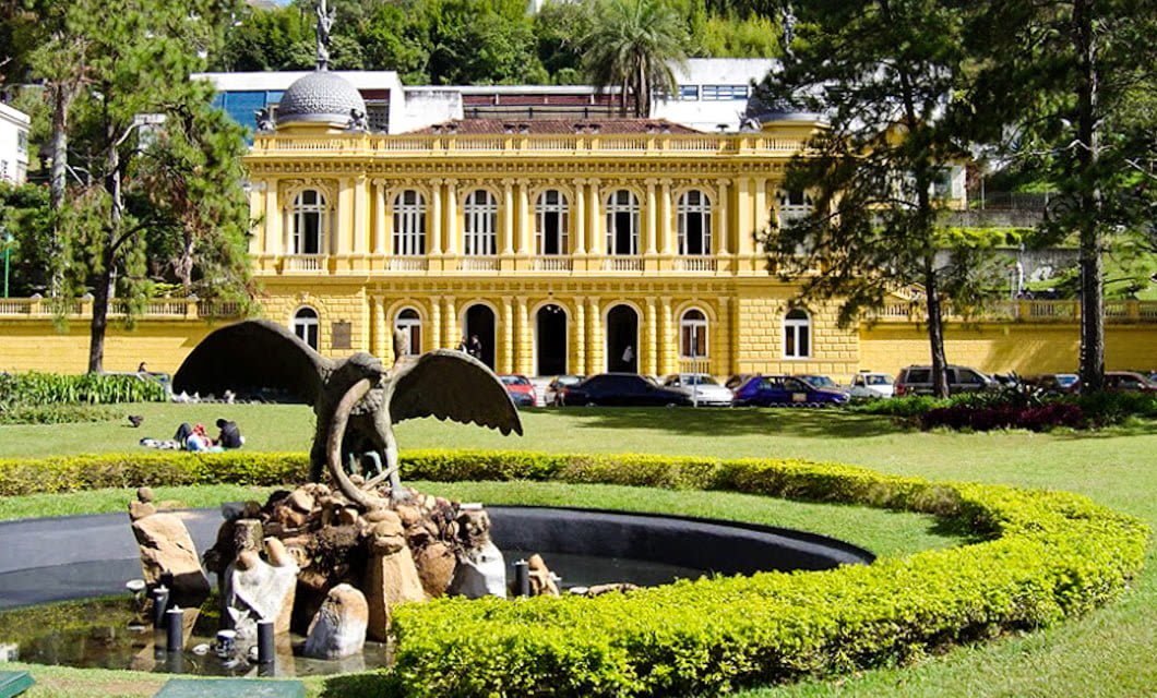 A yellow neoclassical building stands behind a fountain featuring a winged dragon statue in a circular pond, surrounded by green lawns, trees, and people sitting on the grass—one of Rio Tour’s colonial treasures.