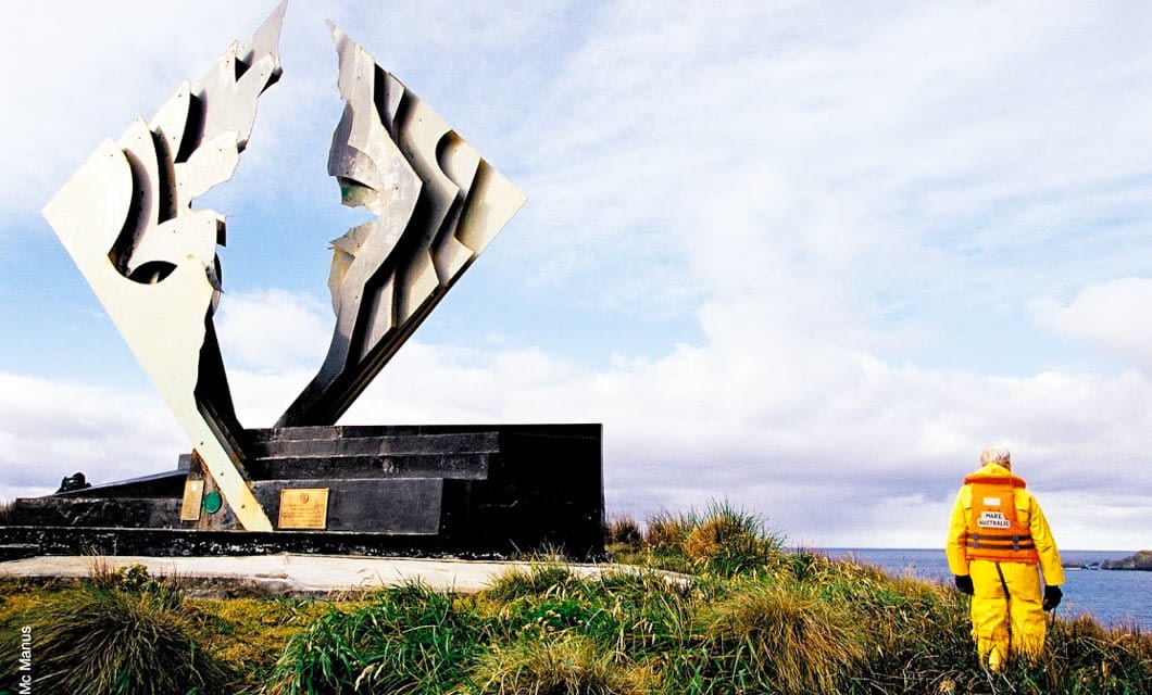 A person in an orange jacket stands near a large, abstract metal sculpture on a dark stone base, set on a grassy area overlooking the ocean—a scene reminiscent of stops along a Patagonia cruise from Santiago to Buenos Aires aboard Mare Australis.