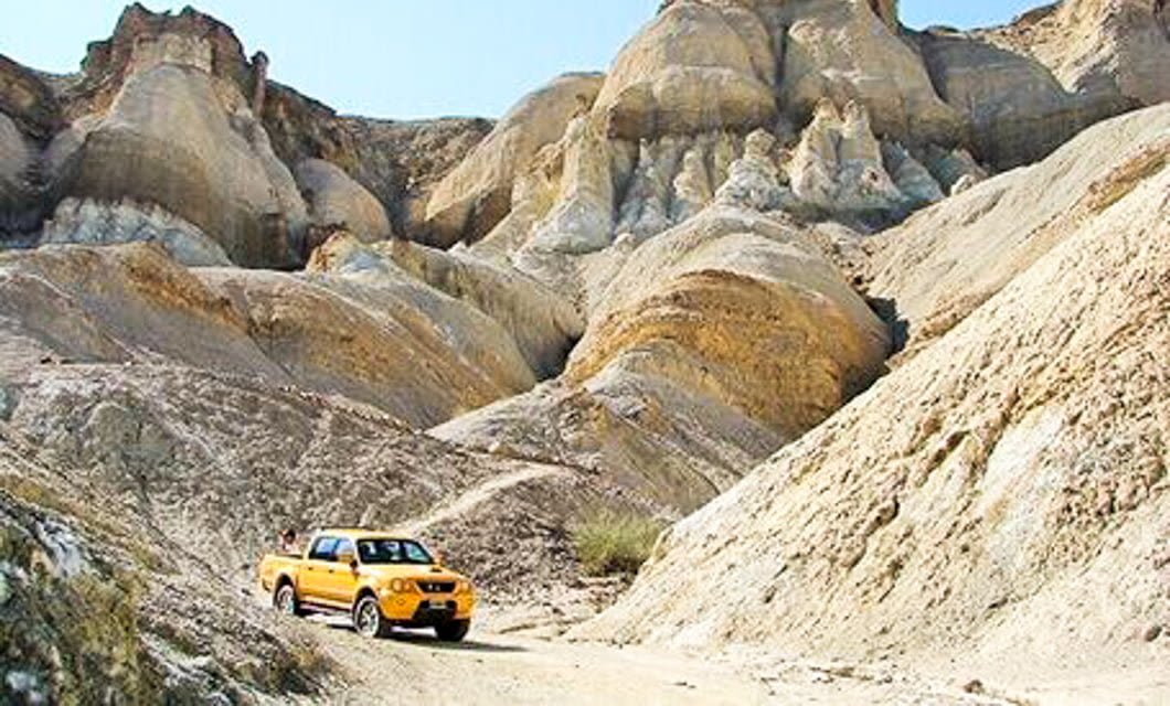 A yellow pickup truck travels on a dirt road, crossing the rugged hills and steep, barren cliffs—its journey unfolding beneath a clear blue sky, as if tracing the Boundaries of Time.