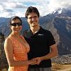 A smiling couple stands close together outdoors, holding hands during their trip, with mountain peaks and a partly cloudy sky in the background. The woman wears sunglasses and an orange dress; the man wears a black shirt.