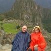 Two people wearing rain ponchos sit and smile in front of ancient stone ruins at Machu Picchu, dreaming of travel arrangements to other parts of the world, with lush green terraces and a mountain peak in the background.