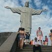 People stand and sit on steps in front of the Christ the Redeemer statue in Rio de Janeiro, Brazil, under a partly cloudy sky, reminiscent of crowds seen at other landmarks like Machu Picchu.