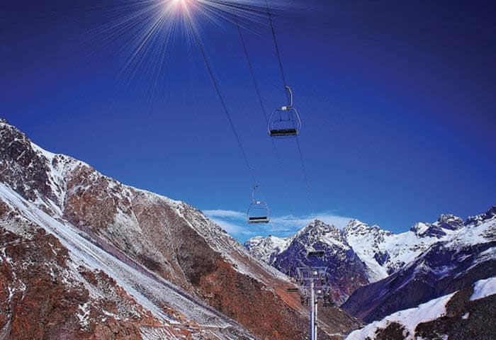 Empty ski lift chairs travel above snowy, rugged mountains under a clear blue sky with the sun shining brightly—a breathtaking scene among the top things to do in Argentina.