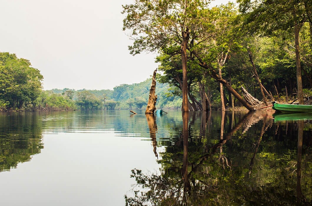A calm river in Bolivia surrounded by lush green trees, their branches leaning over the water and reflecting on its smooth surface. A small green boat is partially visible on the right—one of many tranquil Bolivia attractions to discover.