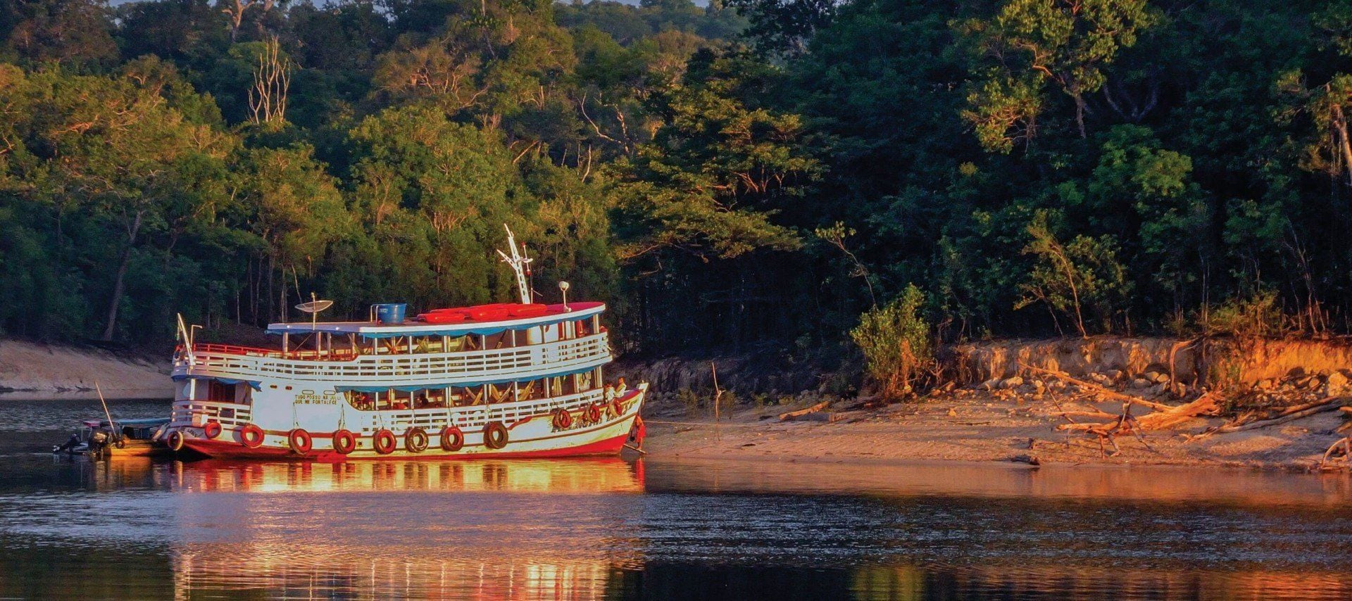 A colorful riverboat with red trim is docked on a sandy riverbank, surrounded by lush green forest under warm sunlight—an inviting scene reminiscent of South America Cruises.