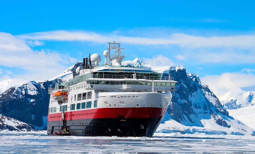 A large red, black, and white cruise ship is docked in icy waters, surrounded by snow-covered mountains and glaciers under a bright blue sky—a perfect scene for unforgettable Antarctica activities and attractions.