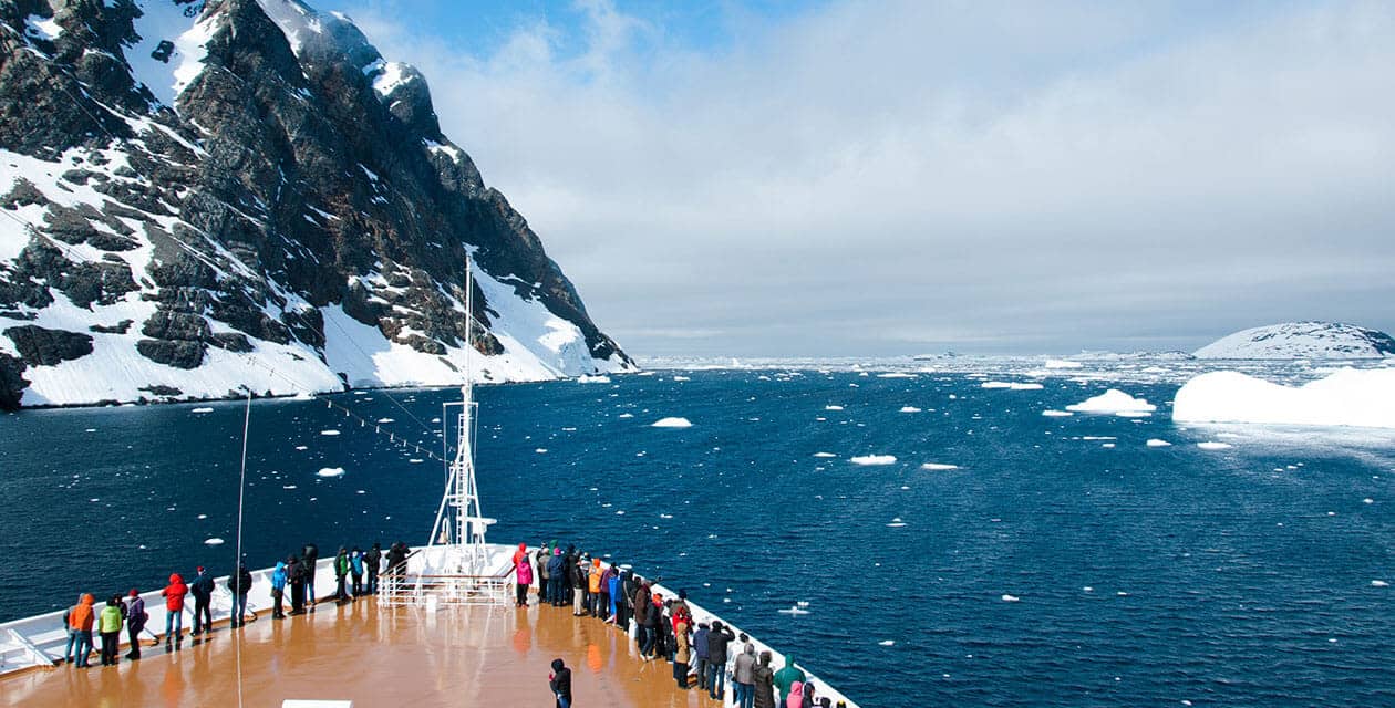 A group of people stand on the bow of a ship, observing icy blue water with floating icebergs and snow-covered mountains under a partly cloudy sky.