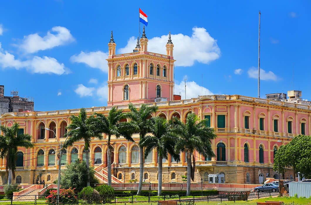 A large, ornate pink and yellow government building with arches and towers stands under a blue sky. The Paraguayan flag flies atop the roof, and palm trees line the front of the building.