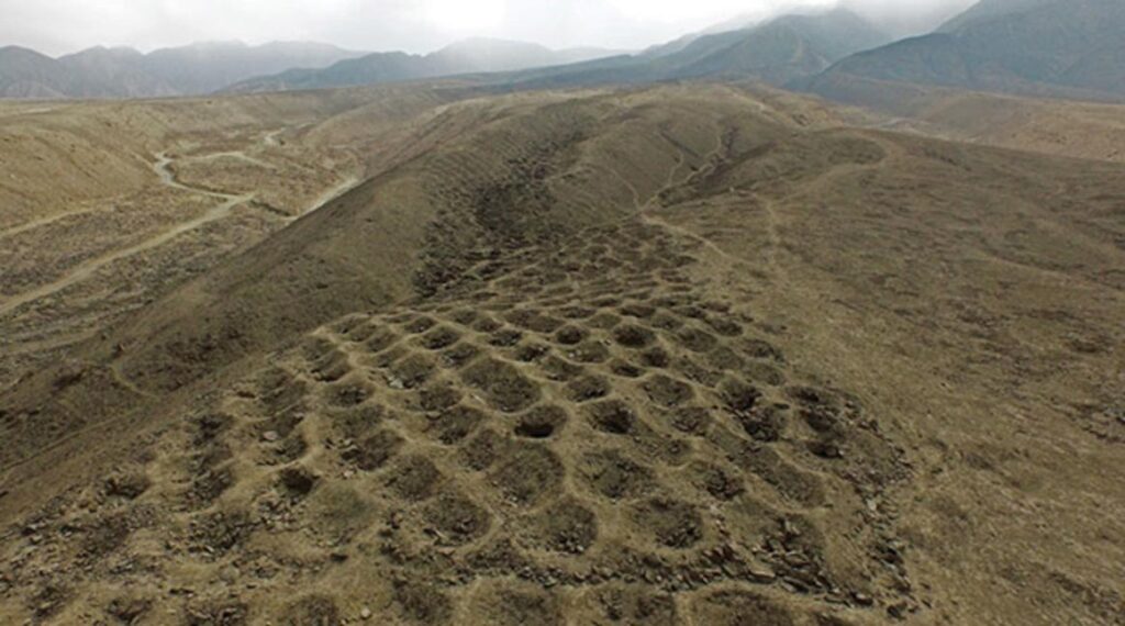 Aerial view of the mysterious site Peru known as the Band of Holes, featuring rows of circular, pit-like holes arranged in a grid pattern along a dry, hilly ridge with mountains in the background under a cloudy sky.