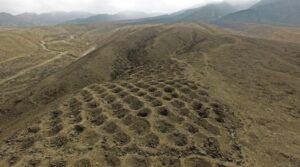 Aerial view of the mysterious site Peru known as the Band of Holes, featuring rows of circular, pit-like holes arranged in a grid pattern along a dry, hilly ridge with mountains in the background under a cloudy sky.