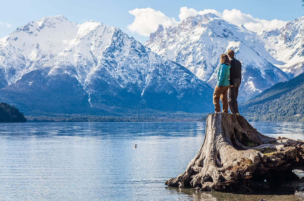 A couple stands on a large tree stump by a calm lake in Patagonia, admiring snow-capped mountains under a blue sky with scattered clouds. Dense green forests line the shore, offering endless things to do for nature lovers and travel enthusiasts alike.