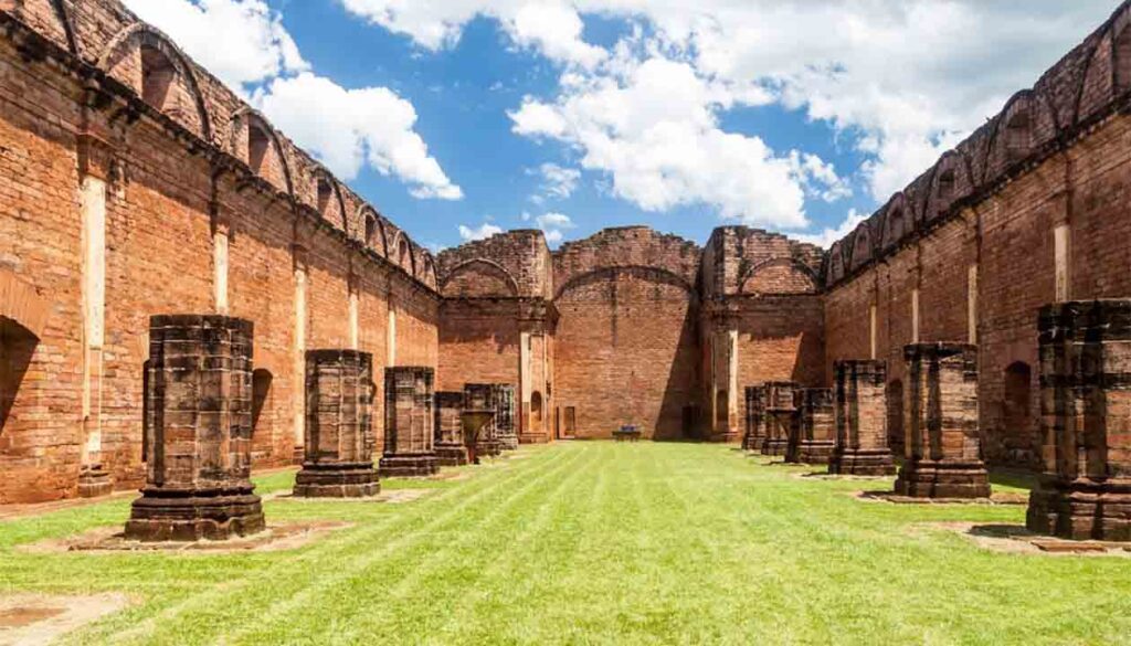 Ruins of a large brick building, with tall walls and partial columns lining a grassy courtyard under a blue sky with clouds—one of the Best Places in Paraguay for history lovers and those looking to Visit Paraguay.