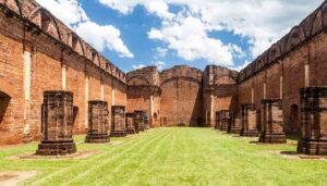 Ruins of a large brick building, with tall walls and partial columns lining a grassy courtyard under a blue sky with clouds—one of the Best Places in Paraguay for history lovers and those looking to Visit Paraguay.
