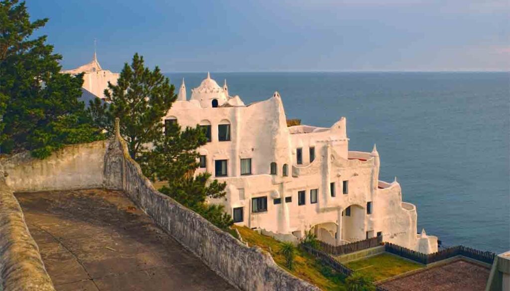 This white, irregularly shaped building with organic, wavy lines overlooks the ocean in South America—one of the best honeymoon destinations—surrounded by greenery and a stone path under a partly cloudy sky.