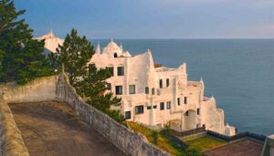 This white, irregularly shaped building with organic, wavy lines overlooks the ocean in South America—one of the best honeymoon destinations—surrounded by greenery and a stone path under a partly cloudy sky.
