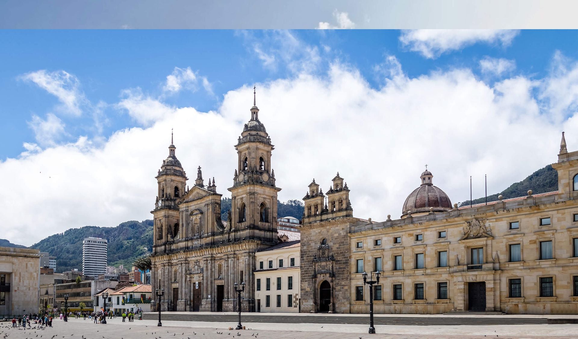A wide view of Bogotá’s Plaza de Bolívar, featuring the neoclassical Primatial Cathedral and historic buildings under a partly cloudy blue sky, with mountains in the background—an essential stop for Colombia travel and exploring things to do in Colombia.