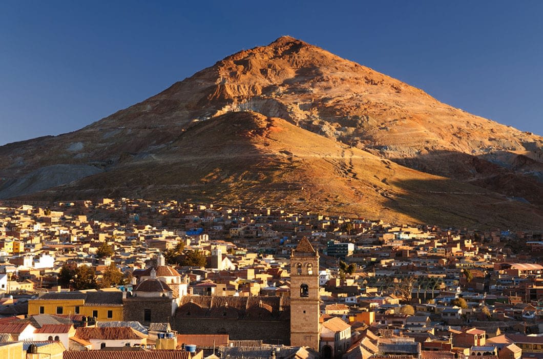 A city with tan and brown buildings sits at the base of a large, sunlit mountain under a clear blue sky. A church with a bell tower is visible among the rooftops—a classic sight among Bolivia attractions for travelers.