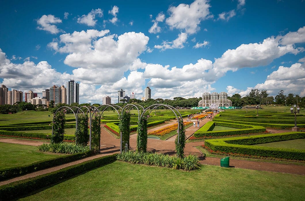 A landscaped garden with trimmed hedges, geometric pathways, green arches, and a glass conservatory in the distance—one of the top Brazil attractions—set against a city skyline under a blue sky with fluffy clouds.