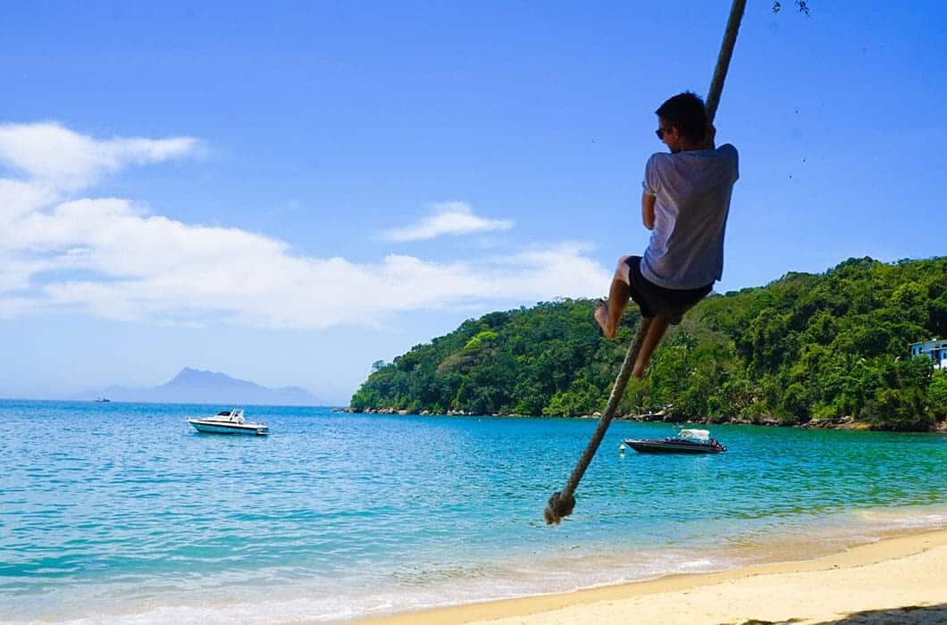 A person swings on a rope over a sandy beach with turquoise water, one of the top Brazil attractions. Two boats float nearby as green hills rise in the background under a bright blue sky with scattered clouds.